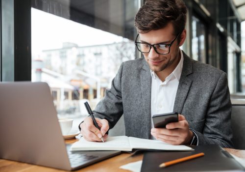 smiling-businessman-eyeglasses-sitting-by-table-cafe-with-laptop-computer-while-using-smartphone-writing-something